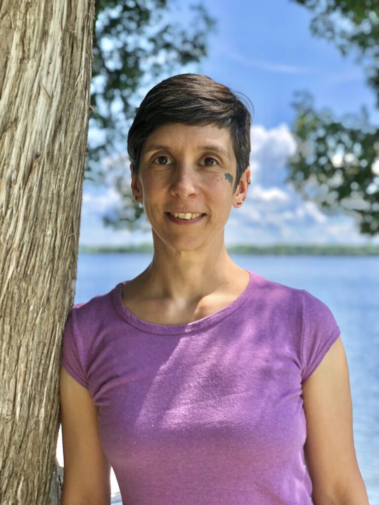 This is a headshot of Arria, leaning against a tree with sky and lake in background