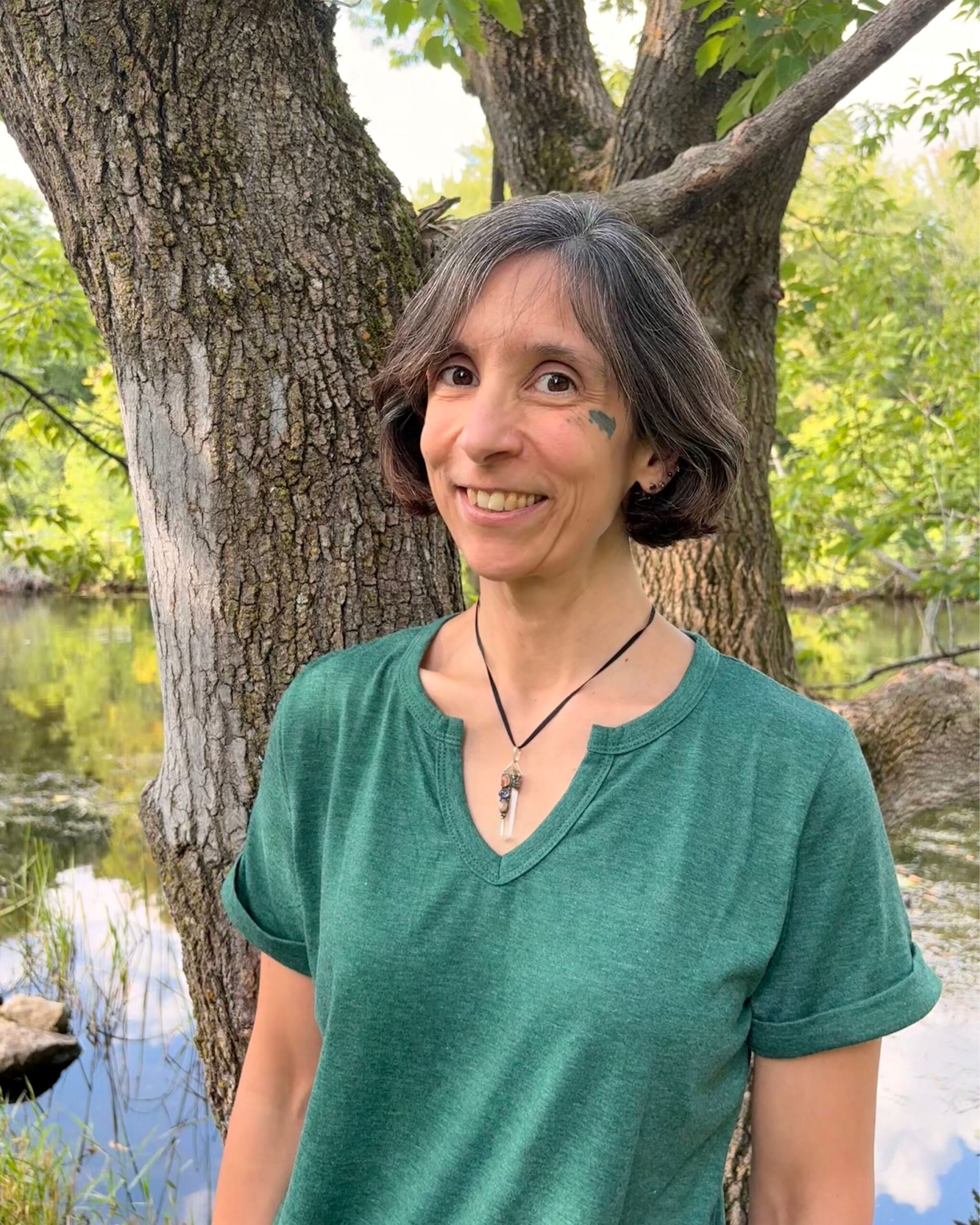 Headshot of Arria from the waist up, smiling, standing by a tree near a river